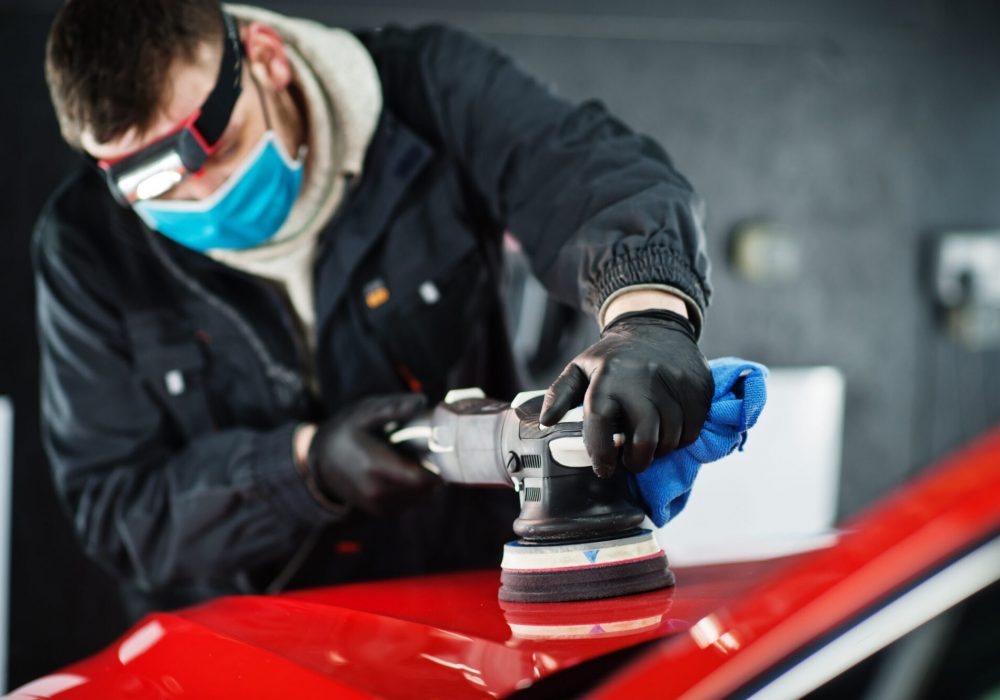 Car detailing concept. Man in face mask with orbital polisher in repair shop polishing orange suv car.
