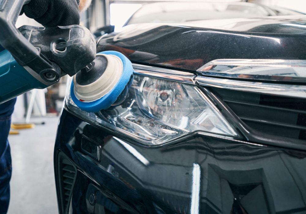Car mechanic pressing polisher instrument with blue pad to front light of automobile during its polishing
